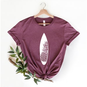 May include: A maroon t-shirt with a white graphic of a surfboard with a pineapple design.