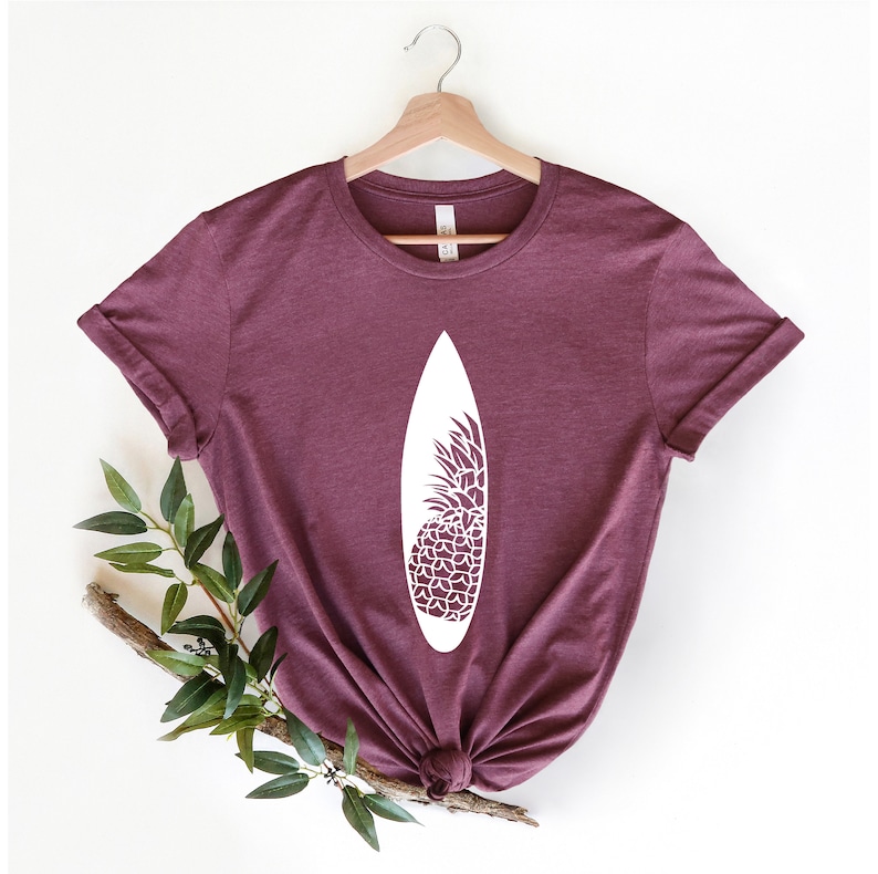 May include: A maroon t-shirt with a white graphic of a surfboard with a pineapple design.