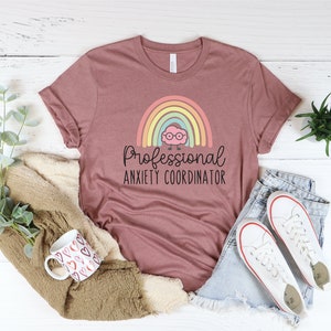 May include: Mauve t-shirt with the text "Professional Anxiety Coordinator" and a cartoon rainbow and brain graphic. The shirt is paired with denim shorts and white sneakers. A mug with heart designs and a beige sweater are also in the image.