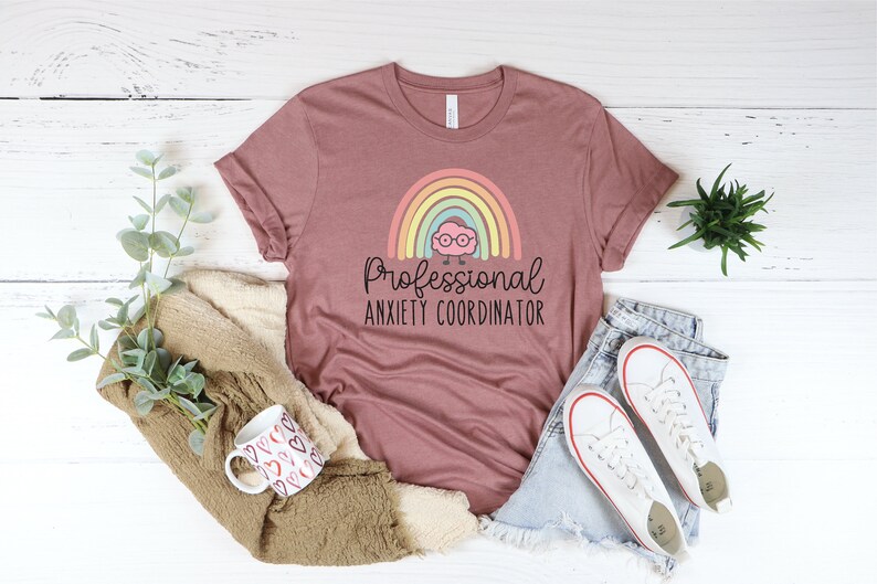 May include: Mauve t-shirt with the text "Professional Anxiety Coordinator" and a cartoon rainbow and brain graphic. The shirt is paired with denim shorts and white sneakers. A mug with heart designs and a beige sweater are also in the image.