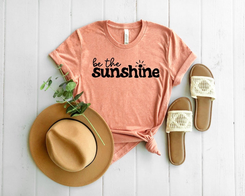 May include: A peach colored t-shirt with the text "be the sunshine" in black lettering with a small sun graphic. The shirt is laid out on a white wooden surface with a brown hat and two white sandals.