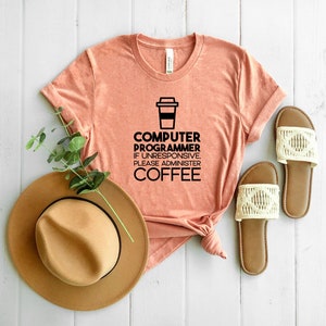 May include: A peach-colored t-shirt with a black graphic of a coffee cup and the text "Computer Programmer. If unresponsive, please administer coffee."