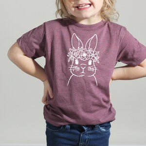 May include: A purple t-shirt with a white outline of a bunny wearing a flower crown.