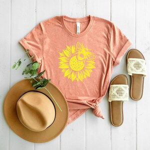 May include: A peach-colored t-shirt featuring a yellow sunflower and bee design. A tan fedora hat and a pair of brown sandals with cream-colored woven straps are also visible. The items are arranged on a white wooden surface.