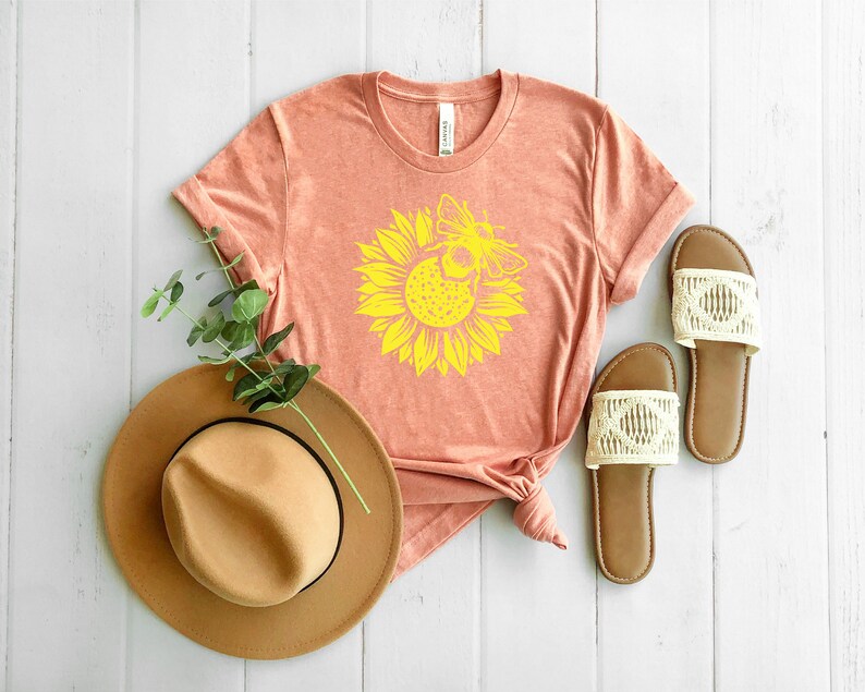 May include: A peach-colored t-shirt featuring a yellow sunflower and bee design. A tan fedora hat and a pair of brown sandals with cream-colored woven straps are also visible. The items are arranged on a white wooden surface.