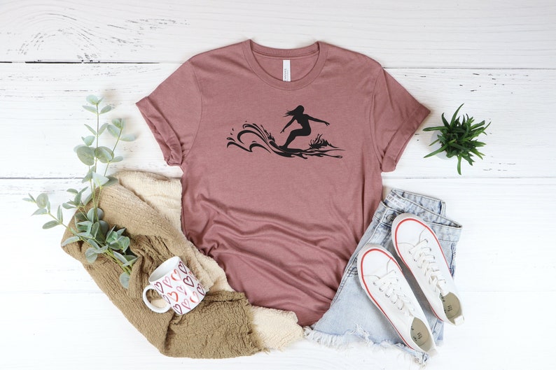 May include: A mauve t-shirt with a black silhouette of a person surfing on a wave.