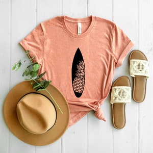 May include: A coral colored t-shirt with a black pineapple design on a surfboard. The shirt is laying on a white wooden surface with a brown hat and white sandals.
