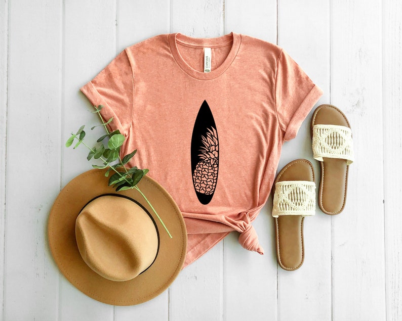 May include: A coral colored t-shirt with a black pineapple design on a surfboard. The shirt is laying on a white wooden surface with a brown hat and white sandals.