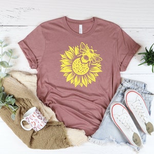 May include: Mauve-colored t-shirt featuring a large yellow sunflower with a bee graphic. The shirt is styled with denim shorts and white sneakers. A mug with heart designs and a cozy blanket are also in the image.