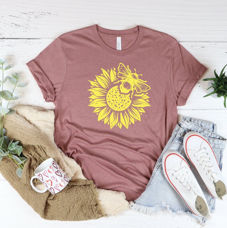 May include: Mauve-colored t-shirt featuring a large yellow sunflower with a bee graphic. The shirt is styled with denim shorts and white sneakers. A mug with heart designs and a cozy blanket are also in the image.