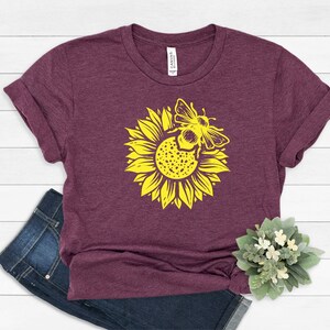 May include: A maroon t-shirt featuring a yellow sunflower with a bee graphic. The shirt has rolled-up sleeves and is paired with blue jeans. A small plant with white flowers is in the bottom right corner. The shirt has the word "CANVAS" printed on the tag.