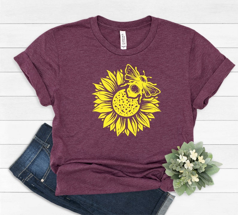 May include: A maroon t-shirt featuring a yellow sunflower with a bee graphic. The shirt has rolled-up sleeves and is paired with blue jeans. A small plant with white flowers is in the bottom right corner. The shirt has the word "CANVAS" printed on the tag.