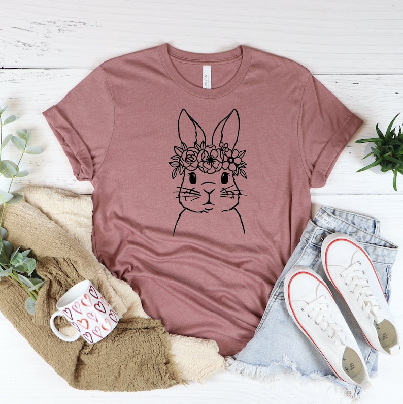 May include: A mauve t-shirt with a black line drawing of a bunny wearing a flower crown. The bunny is looking directly at the viewer.