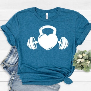 May include: A teal blue t-shirt with a white graphic of a heart shaped kettlebell with dumbbells on either side.