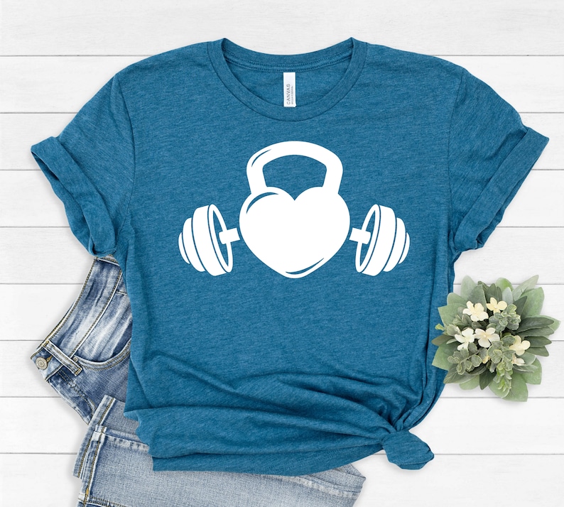 May include: A teal blue t-shirt with a white graphic of a heart shaped kettlebell with dumbbells on either side.