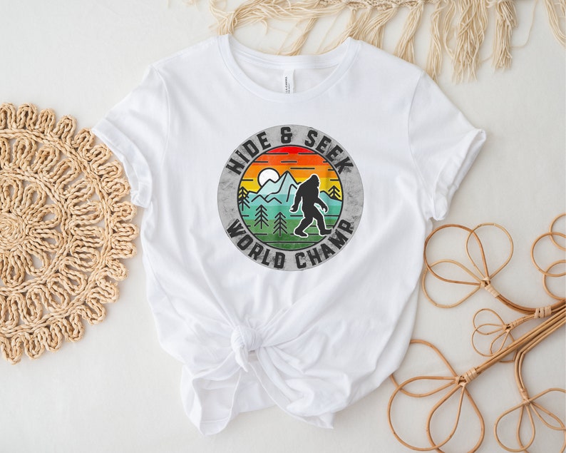 May include: White t-shirt with a vintage-style graphic of a Bigfoot walking in a forest with the text "Hide & Seek World Champ".