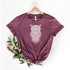 May include: A maroon t-shirt with a white, detailed owl design on the front. The owl is stylized with intricate patterns and a mandala-like design.