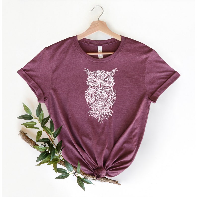 May include: A maroon t-shirt with a white, detailed owl design on the front. The owl is stylized with intricate patterns and a mandala-like design.