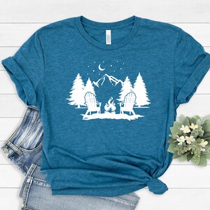 Pu&ograve; includere: Una maglietta blu turchese con un design grafico bianco di due sedie di fronte a un fal&ograve; davanti a una catena montuosa con alberi e una luna crescente nel cielo.