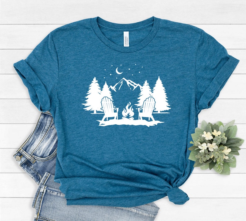 Pu&ograve; includere: Una maglietta blu turchese con un design grafico bianco di due sedie di fronte a un fal&ograve; davanti a una catena montuosa con alberi e una luna crescente nel cielo.