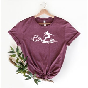 May include: A maroon t-shirt with a white silhouette of a person surfing a wave.