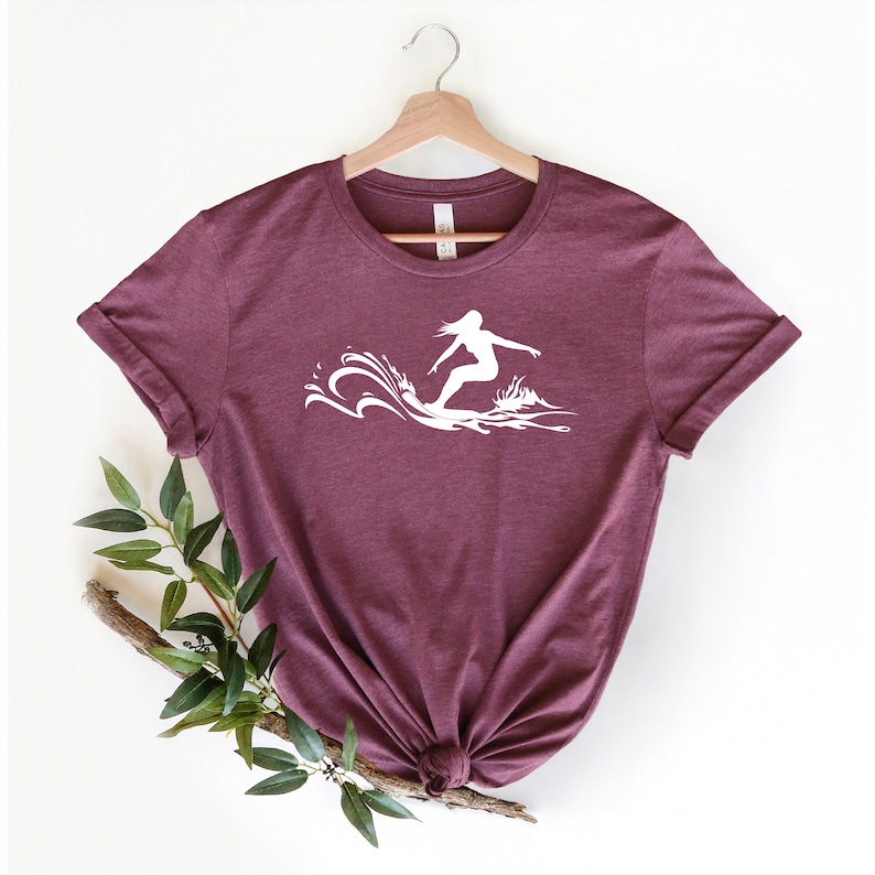 May include: A maroon t-shirt with a white silhouette of a person surfing a wave.