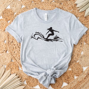May include: A gray t-shirt with a black silhouette of a person surfing on a wave.