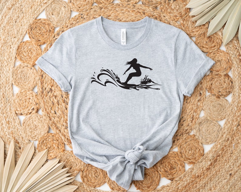 May include: A gray t-shirt with a black silhouette of a person surfing on a wave.