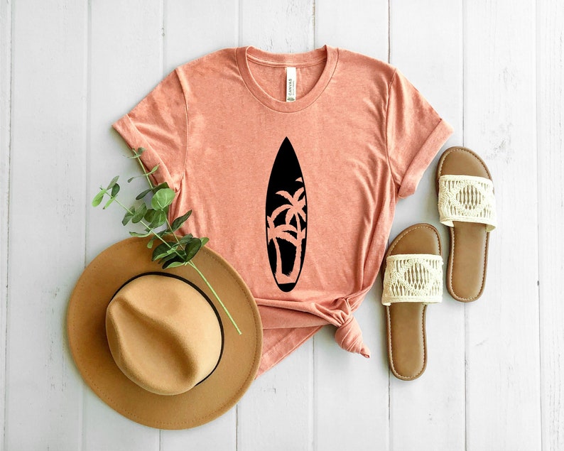 May include: A coral colored t-shirt with a black silhouette of a surfboard with palm trees on it. The shirt is laid out on a white wooden surface with a brown hat and two white sandals.