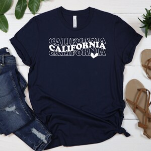 Puede incluir: Una camiseta azul marino con el texto "CALIFORNIA" repetido tres veces en blanco, con un s&iacute;mbolo de coraz&oacute;n al final de la &uacute;ltima palabra.