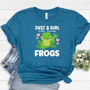 K&ouml;nnte beinhalten: Ein blaues T-Shirt mit dem Text "Just a girl who loves frogs" und einem Cartoon-Frosch mit rosa Herzen.