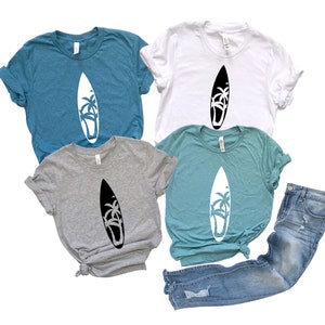 May include: Four t-shirts with a graphic of a surfboard with palm trees on it. The shirts are in different colors: teal, white, gray, and light blue. The shirts are laid out on a white background.
