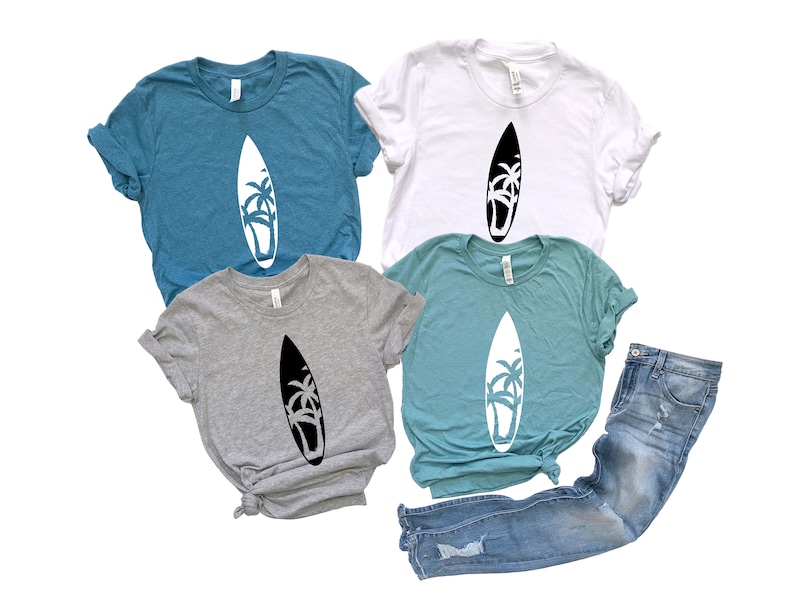 May include: Four t-shirts with a graphic of a surfboard with palm trees on it. The shirts are in different colors: teal, white, gray, and light blue. The shirts are laid out on a white background.