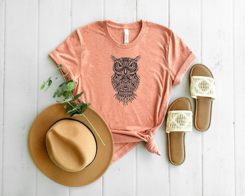 May include: A peach-colored t-shirt with a black and white mandala-style owl graphic. The shirt is made of a soft, comfortable material and has a relaxed fit.