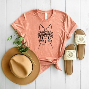 May include: A peach-colored t-shirt with a black and white illustration of a bunny wearing a flower crown. The t-shirt is laid out on a white wooden surface with a brown hat and white sandals.
