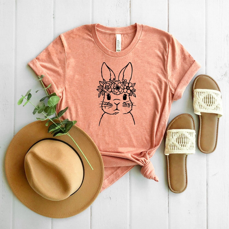 May include: A peach-colored t-shirt with a black and white illustration of a bunny wearing a flower crown. The t-shirt is laid out on a white wooden surface with a brown hat and white sandals.