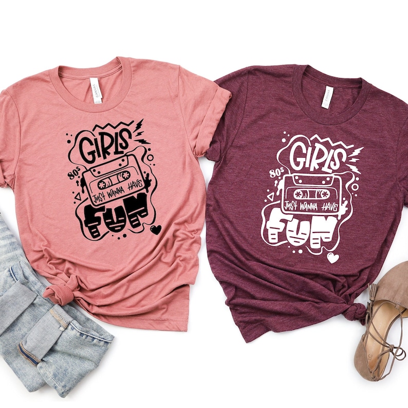 K&ouml;nnte beinhalten: Zwei T-Shirts mit einem grafischen Design eines Kassettenbandes mit dem Text "Girls Just Wanna Have Fun" und "80s" auf einem rosa und maroonfarbenen Shirt.