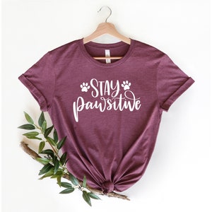 May include: Burgundy t-shirt with white text that reads "Stay Pawsitive" with paw prints on either side of the word "Stay".