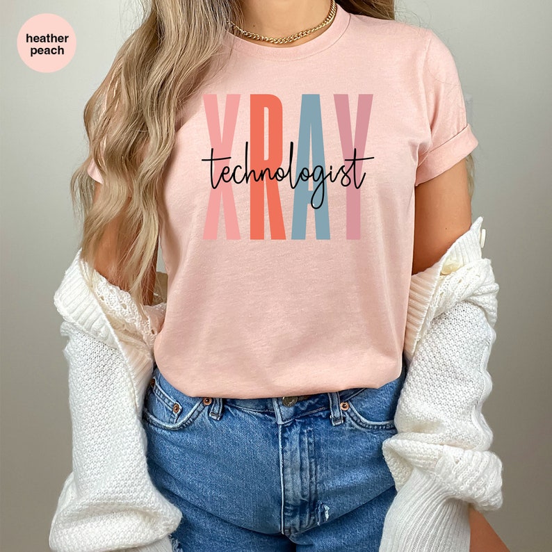 May include: A heather peach t-shirt with the text "XRAY technologist" in colorful letters. The letters are in shades of pink, orange, blue, and light pink.