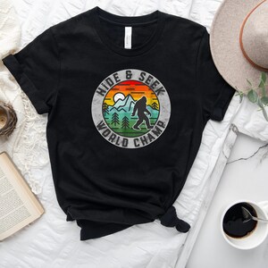 May include: Black t-shirt with a vintage-style graphic of a Bigfoot in a forest scene. The graphic says "Hide & Seek World Champ".