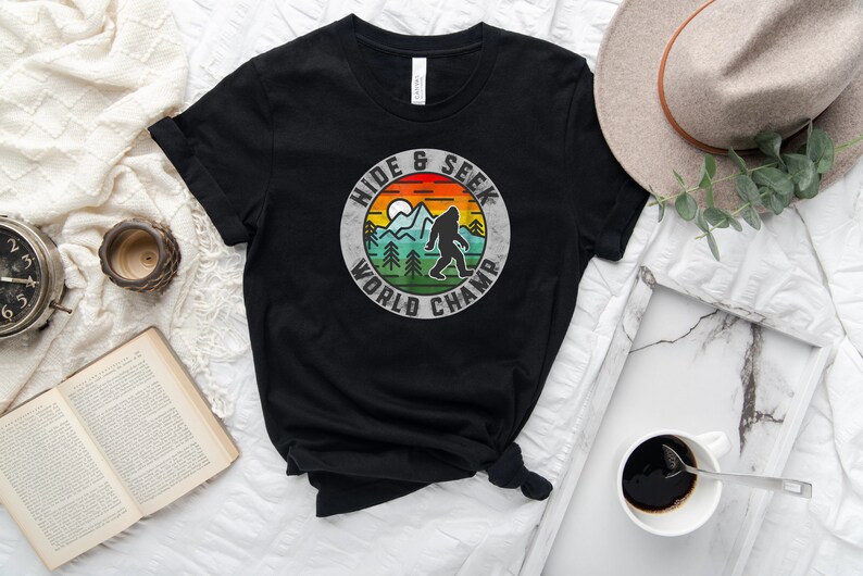 May include: Black t-shirt with a vintage-style graphic of a Bigfoot in a forest scene. The graphic says "Hide & Seek World Champ".