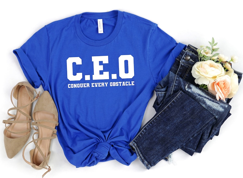 May include: A royal blue t-shirt with white text that reads "C.E.O. CONQUER EVERY OBSTACLE". The shirt is folded and tied in a knot. A pair of brown sandals and a pair of blue jeans are also in the image.