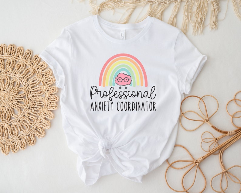 May include: White t-shirt featuring a colorful rainbow design with a cartoon brain wearing glasses. The text "Professional Anxiety Coordinator" is printed below the rainbow. The shirt is made of a soft material.