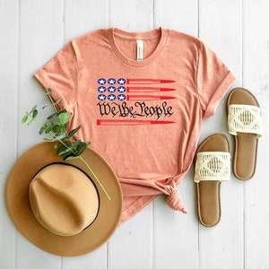 May include: A peach-colored t-shirt with a red, white, and blue American flag design and the text "We the People" printed on the front. The flag design features five red bullets.