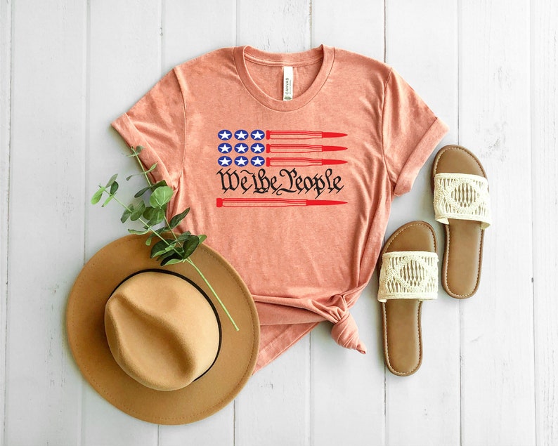 May include: A peach-colored t-shirt with a red, white, and blue American flag design and the text "We the People" printed on the front. The flag design features five red bullets.