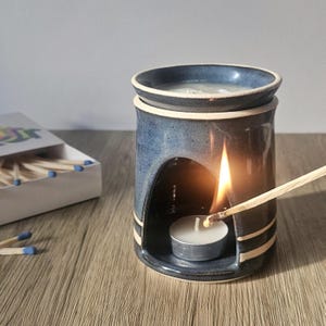 May include: A blue ceramic essential oil diffuser with a lit tea light candle inside. A match is being used to light the candle. The diffuser has a round top and a curved opening. A box of matches is in the background.