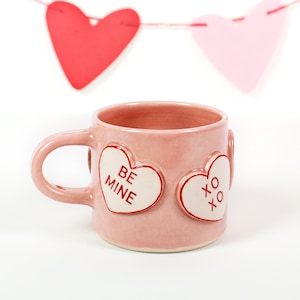 May include: A pink ceramic mug with a handle, featuring two heart-shaped embellishments. One heart reads "BE MINE" and the other says "XO XO" in red text. A garland of red and pink paper hearts hangs in the background.
