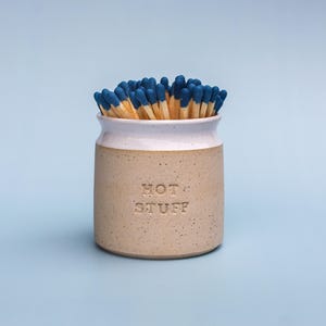 May include: A beige ceramic match holder with the words "HOT STUFF" embossed on the front. The holder is filled with wooden matches with blue tips. The rim of the holder is white, set against a light blue background.