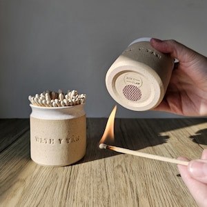 May include: A ceramic match holder and striker set. The beige-colored holder has a white rim and the text "WITH Y TAN" on the side. The bottom of the matching striker has the text "ZOES - CLAY" and a red striker pad. A match is lit.