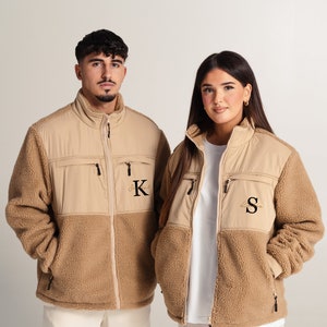 May include: Two people wearing brown fleece jackets with a contrasting beige panel. The jackets have zipper closures and pockets. The jacket on the left has a "K" embroidered on the chest, and the jacket on the right has an "S" embroidered on the chest.